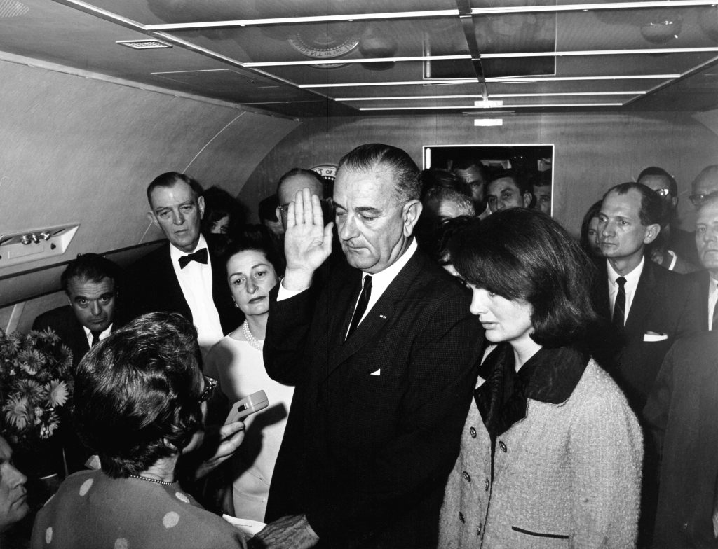 President Lyndon B. Johnson takes the oath of office on Air Force One at Love Field in Dallas, Texas, following the assassination of President John F. Kennedy. Left to right: Assistant Press Secretary, Malcolm Kilduff (holding dictating machine, partially out of frame); Judge Sarah T. Hughes (administering oath); Jack Valenti; Congressman Albert Thomas of Texas; Secretary to President Johnson, Marie Fehmer (partially hidden behind Thomas); First Lady Lady Bird Johnson; Dallas Police Chief, Jesse Curry (face hidden by Vice President’s raised hand); President Johnson; Secretary to President Kennedy, Evelyn Lincoln (mostly hidden behind Mrs. Kennedy); Congressman Homer Thornberry of Texas (mostly hidden behind Lincoln); Secret Service agents Roy Kellerman (partially hidden behind Thornberry) and Thomas “Lem” Johns (partially hidden behind Mrs. Kennedy); former First Lady Jacqueline Kennedy; Mrs. Kennedy’s Press Secretary, Pamela Turnure (partially hidden behind Brooks); Congressman Jack Brooks of Texas; deputy director of public affairs for the Peace Corps, Bill Moyers (mostly obscured by Brooks); President Kennedy’s physician, Admiral Dr. George G. Burkley (on edge of frame); others in background.