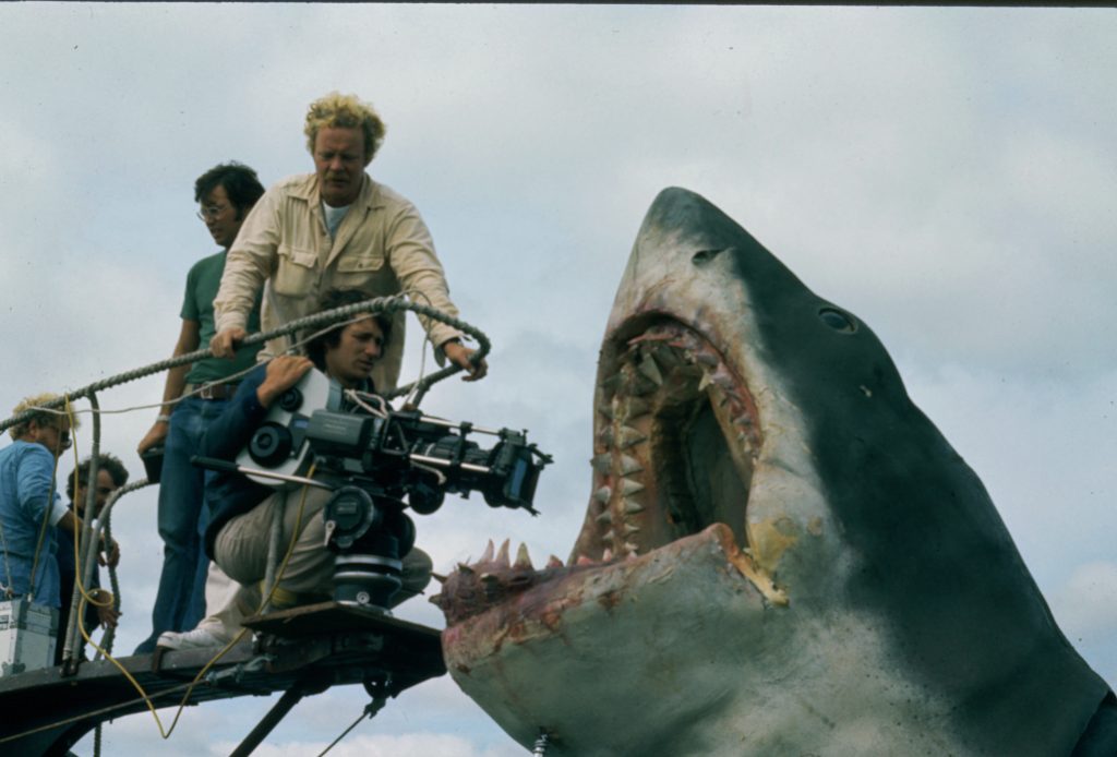 A still from Jaws: The Exhibition: Director Steven Spielberg, kneeling with camera, during production of Jaws (1975). Others unidentified.